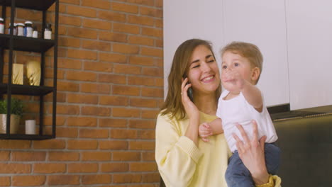 woman holding her baby in the kitchen while talking on the phone