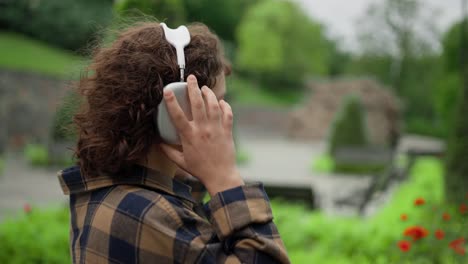 A-brunette-student-girl-with-curly-hair-and-a-plaid-shirt-is-relaxing-in-the-park-and-listening-to-music-on-wireless-headphones