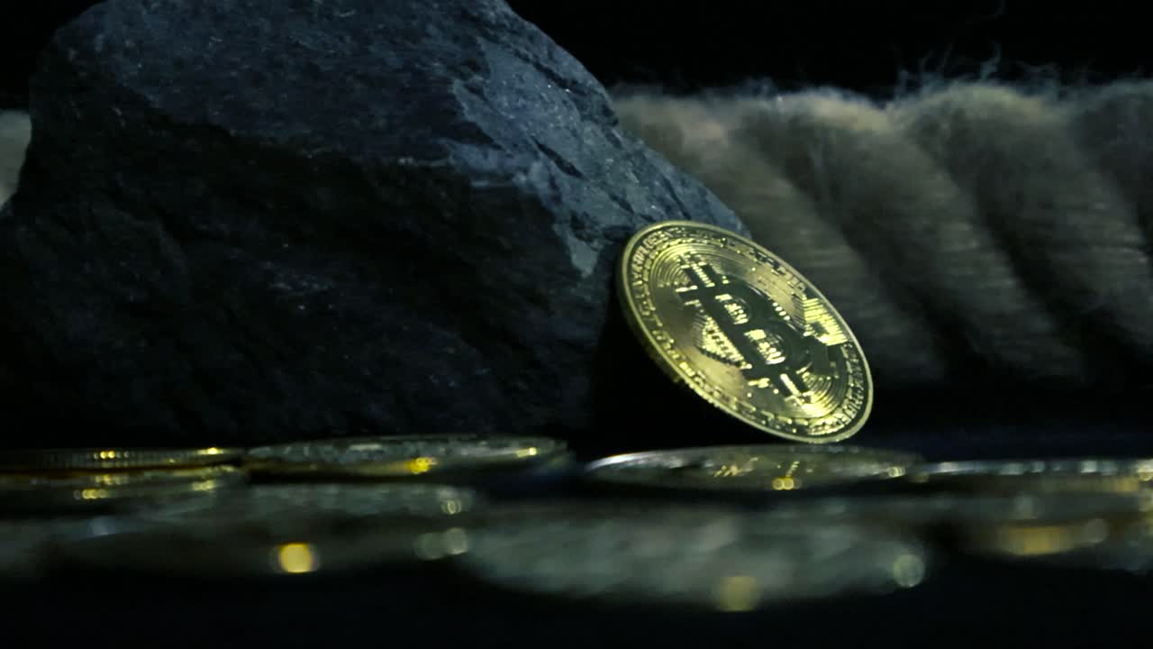 Low angle close up footage of golden yellow reflective bitcoin cryptocurrency coins on a dark studio background with good lighting, a rope and rocks. One bitcoin is standing against the large rock.