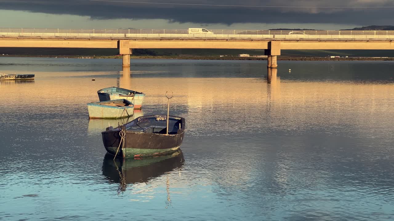 Small fishing boats are gently swaying in the tranquil waters of barbate, spain, as the sun sets, casting a warm glow on the scene while a truck crosses a bridge in the background