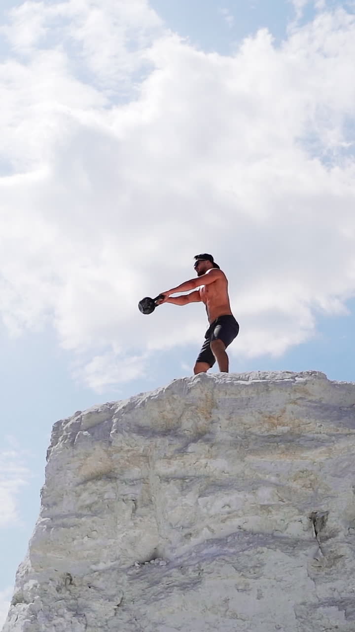 Strong man lifting kettlebell on the mountain in summer. Male athlete swinging kettlebell with two hands on the beautiful natural background outdoors. Vertical video