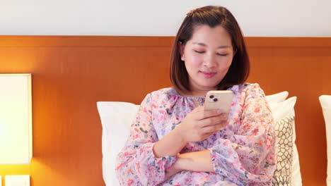 Positive-beautiful-female-holding-a-mobile-phone-sitting-on-the-bed-in-her-bedroom-at-home