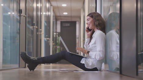 Joven-Empresaria-Sentada-En-El-Suelo-En-El-Pasillo-De-Una-Oficina-Moderna-Trabajando-En-Una-Computadora-Portátil-Hablando-Por-Teléfono-Móvil