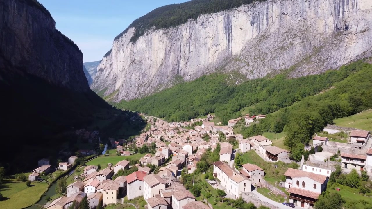 Valle di un villaggio aerea sotto una scogliera imponente, ripresa cinematografica 4K con drone che rivela case in pietra illuminate dal sole, vicolo tortuoso.