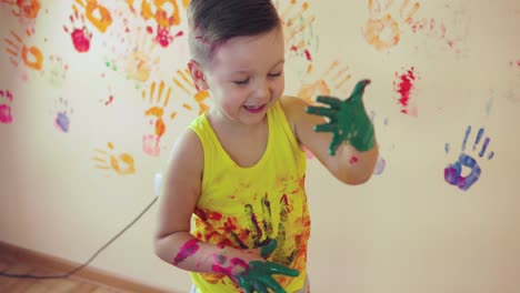 close up veiw of cute little boy with painted hands after leaving his colorful handprints on the wall and showing his thumb up. young happy family. mother and child concept