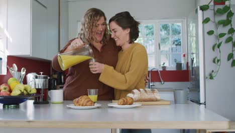 Happy-caucasian-lesbian-couple-embracing-and-pouring-orange-juice-in-kitchen