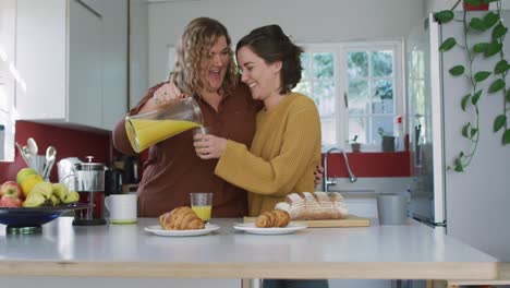 feliz pareja de lesbianas caucásicas abrazándose y vertiendo jugo de naranja en la cocina