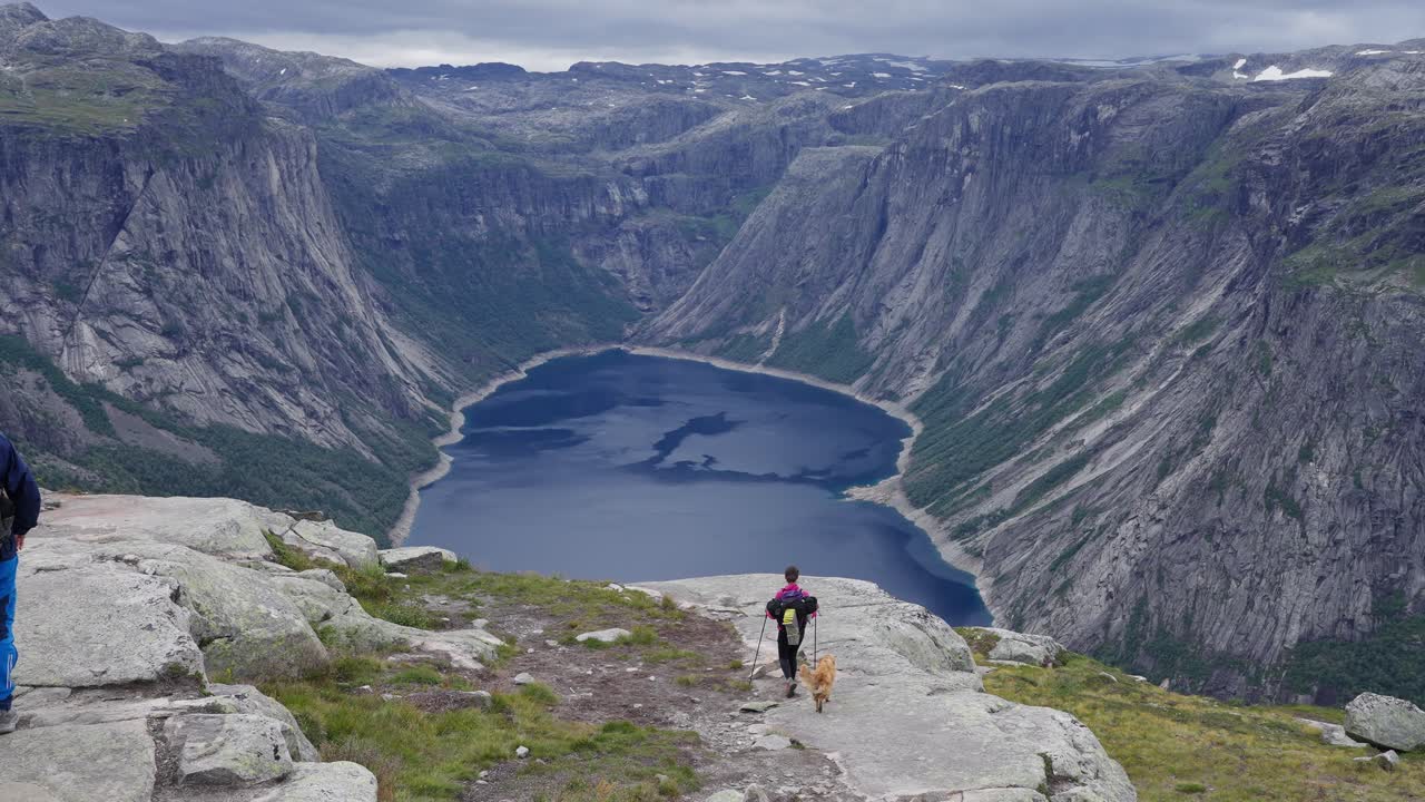 Man and dog hiking to cliff edge with stunning fjord and Norway mountains ahead, Trolltunga