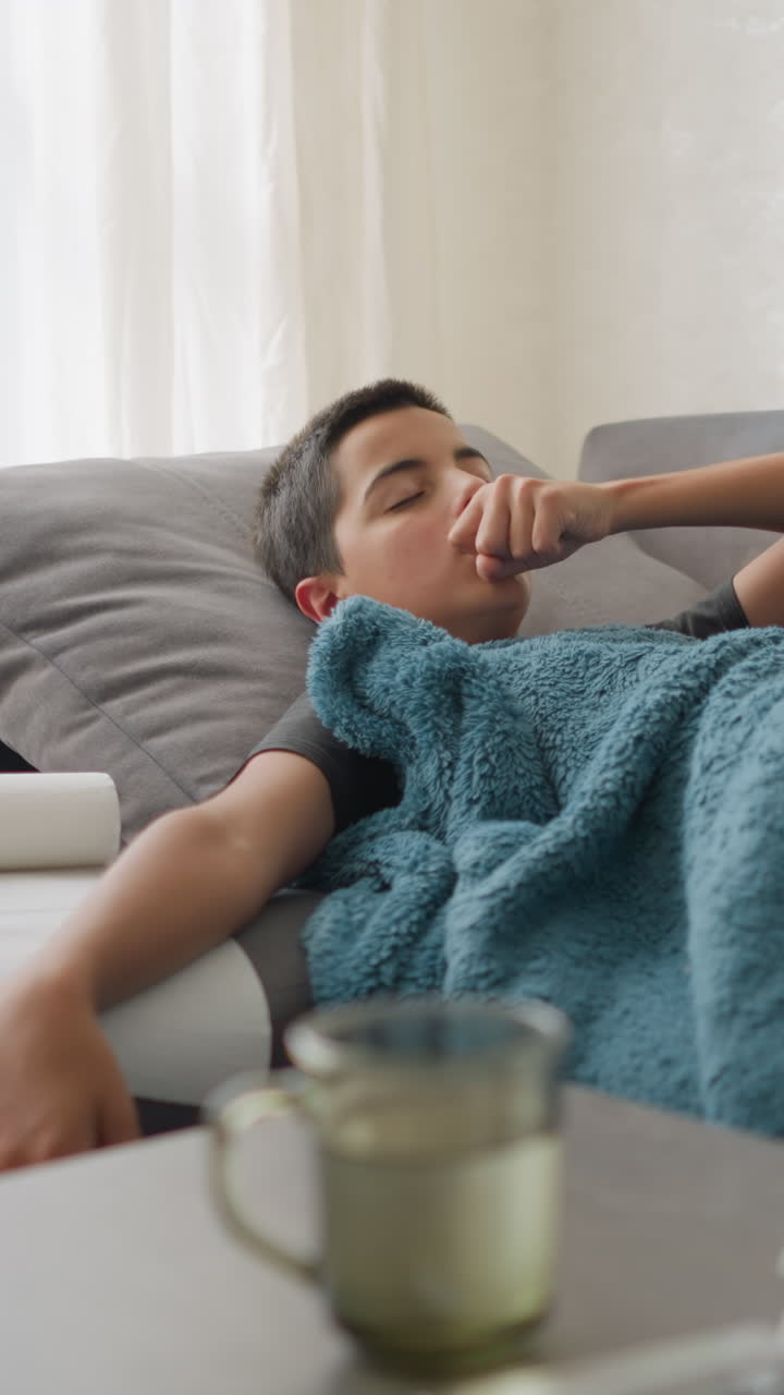 Sick boy resting on couch, hand on forehead, with used tissues, thermometer, and glass cup on the table, boy covered in blanket, recovering from illness in living room