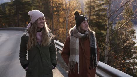 positive female tourists in winter coats walking in slow motion on a long road through the countryside towards the snowy mountain peaks, golden trees and lake on the background. smiling caucasian girls exploring norway nature