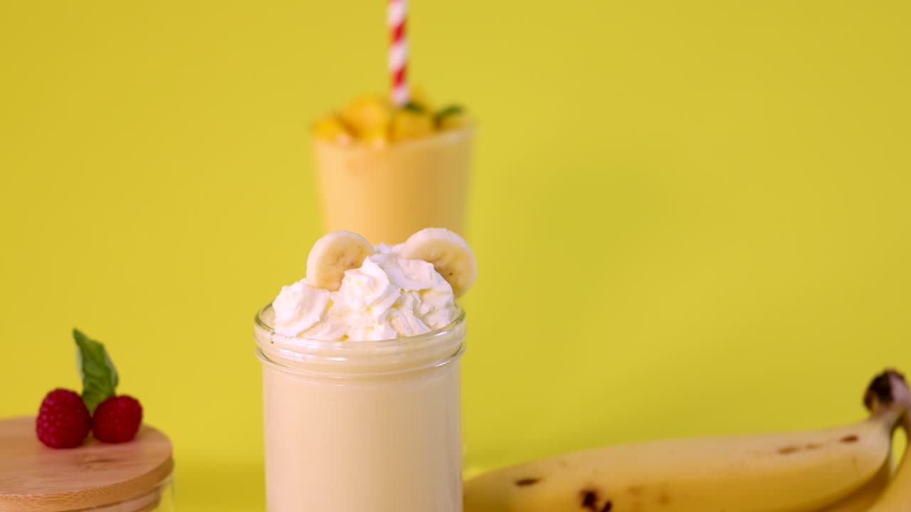 Hand places banana slices on whipped cream-topped banana milkshake, bright yellow background, studio lighting