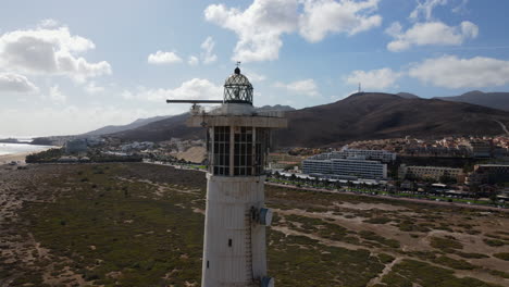 Toma-Aerea-En-Orbita-Ya-Poca-Distancia-Del-Faro-De-Morro-Jable,-Con-Los-Edificios-De-La-Ciudad-Y-La-Playa-De-Morro-Jable-Al-Fondo