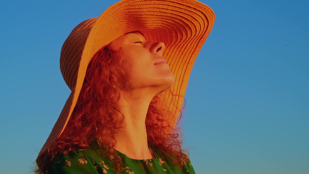 A Joyful Winter Evening: A Young Woman in a Bright Floral Dress and Wide-Brimmed Hat Enjoys a Beautiful Sunset by the Seashore with Radiant Colors