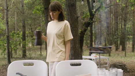 happy woman bringing coffee pot to table to have breakfast with her husband at the camping in the forest