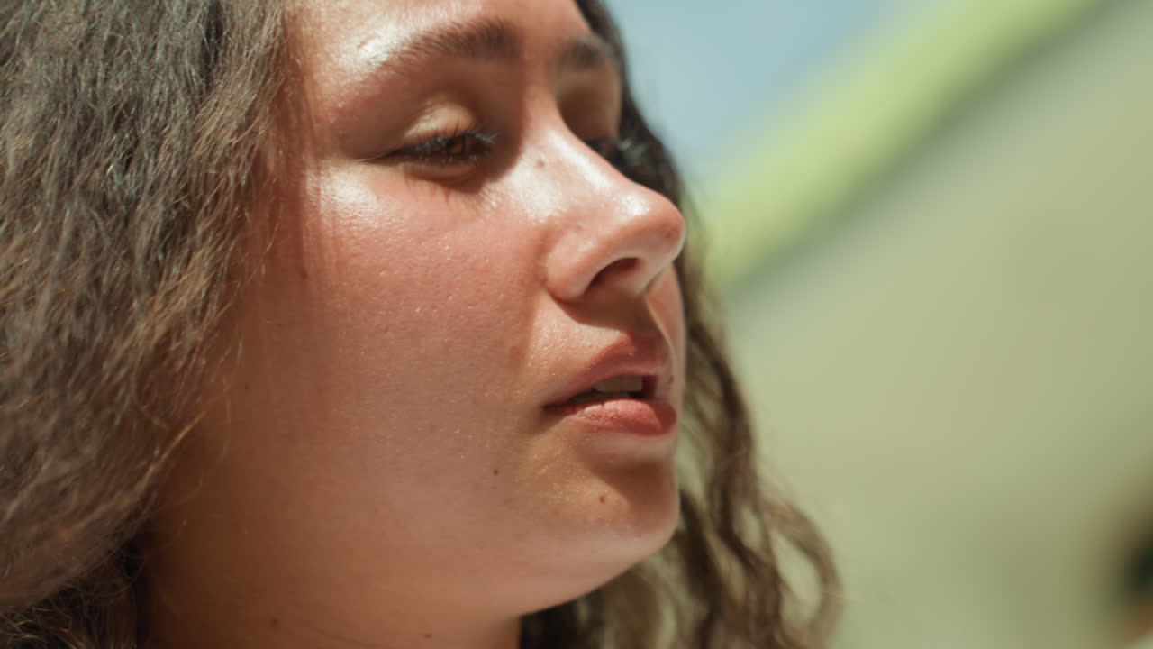 Primo piano di una donna che parla vicino alla riva, un'amica in riva al mare che pronuncia un monologo spontaneo sul lungomare dell'isola, con luce solare soffusa sul viso, labbra e occhi espressivi, gioielli casual e un'atmosfera di conversazione calda.