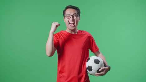 공을 들고 있는 아시아인 남자가 골을 외치며 우승팀을 축하하고 초록색 스크린 배경에서 축구를 응원하고 있다.
