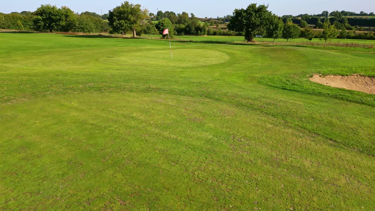 Green grassy and cutted golf fairway with a red flag fluttering in gentle sunlight