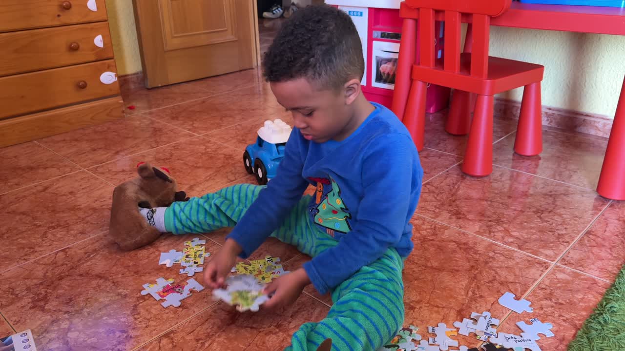 4-year-old black kid sitting on the foor at home doing a puzzle. He is wearing curly hair and a blue winter pyjamas.