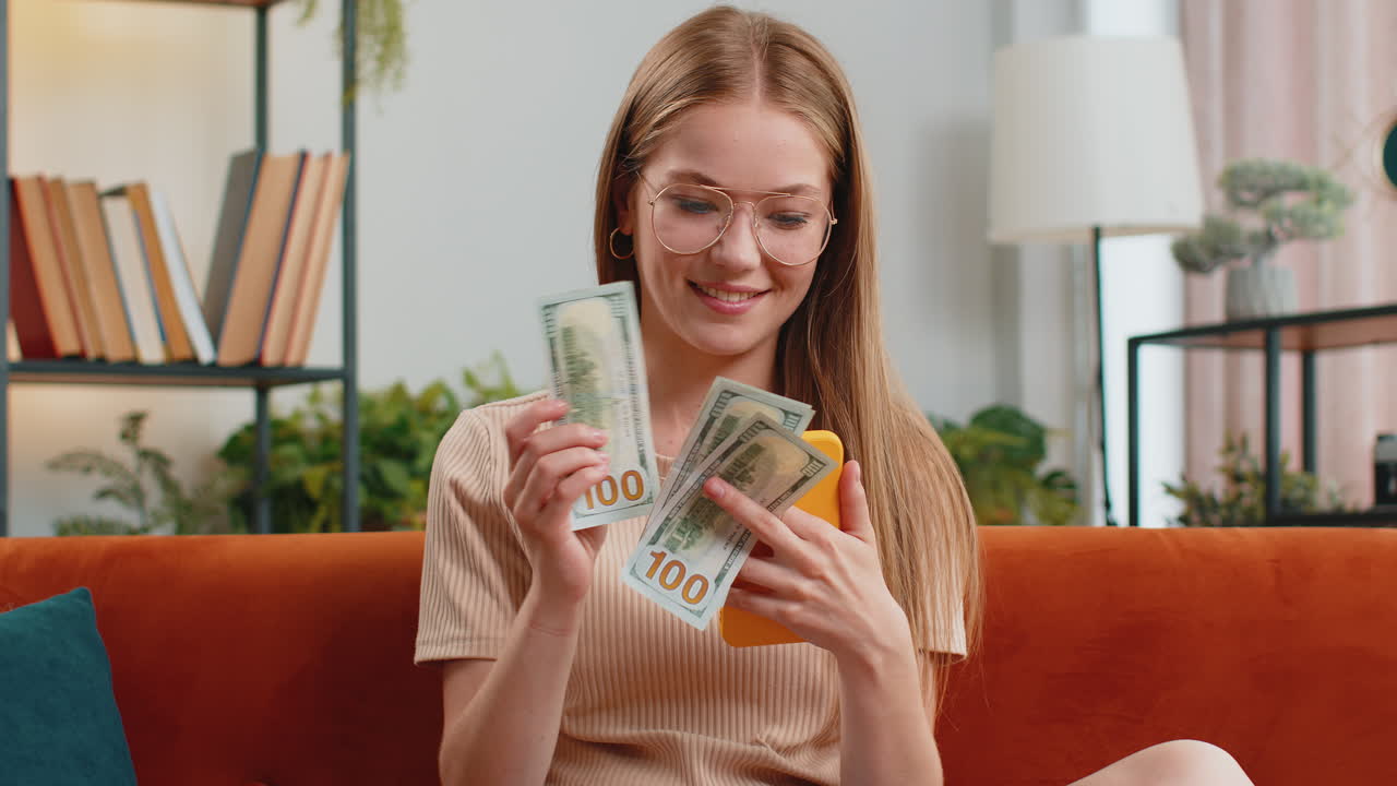 Smiling happy young woman counting money cash use smartphone income saves lottery win budget at home