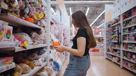 side view of woman holding and inspecting vibrant orange plush purse with green details in well-lit mall, she carefully examines it while browsing shelves, with blurred shoppers walking in background