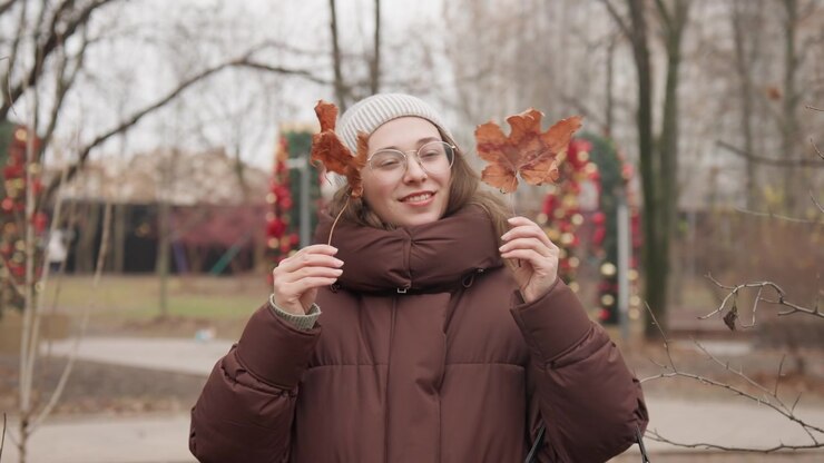 Donna bianca con cappotto marrone che lancia foglie durante una passeggiata in un parco urbano, indossa un berretto grigio e occhiali, un piumino accogliente, porta una borsa nera. Espressione gioiosa, foglie che svolazzano al rallentatore, cielo coperto.