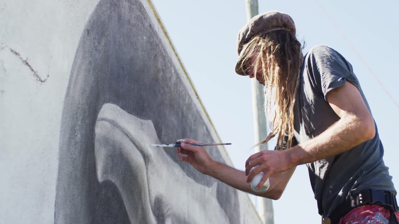 video de un artista caucásico con dreadlocks pintando un mural de ballena en la pared