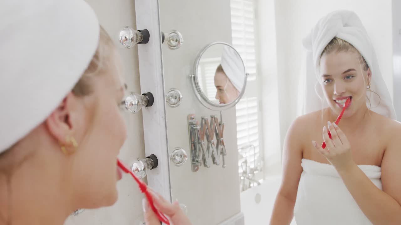 Happy caucasian plus size woman in front of mirror with towel on head brushing teeth in slow motion