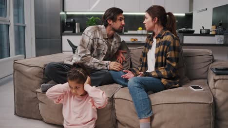 Sad-and-withdrawn-little-brunette-girl-in-a-pink-suit-covers-her-ears-with-her-palms-while-her-parents-in-a-checkered-brunette-shirt-are-arguing-while-sitting-on-the-sofa-in-a-modern-studio-apartment