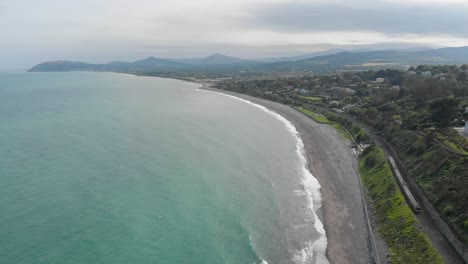 Panoramic-ultrawide-shot-of-train-passing-by-the-beautiful-green-sea-on-a-bright-sunny-day-with-mountains-in-the-background