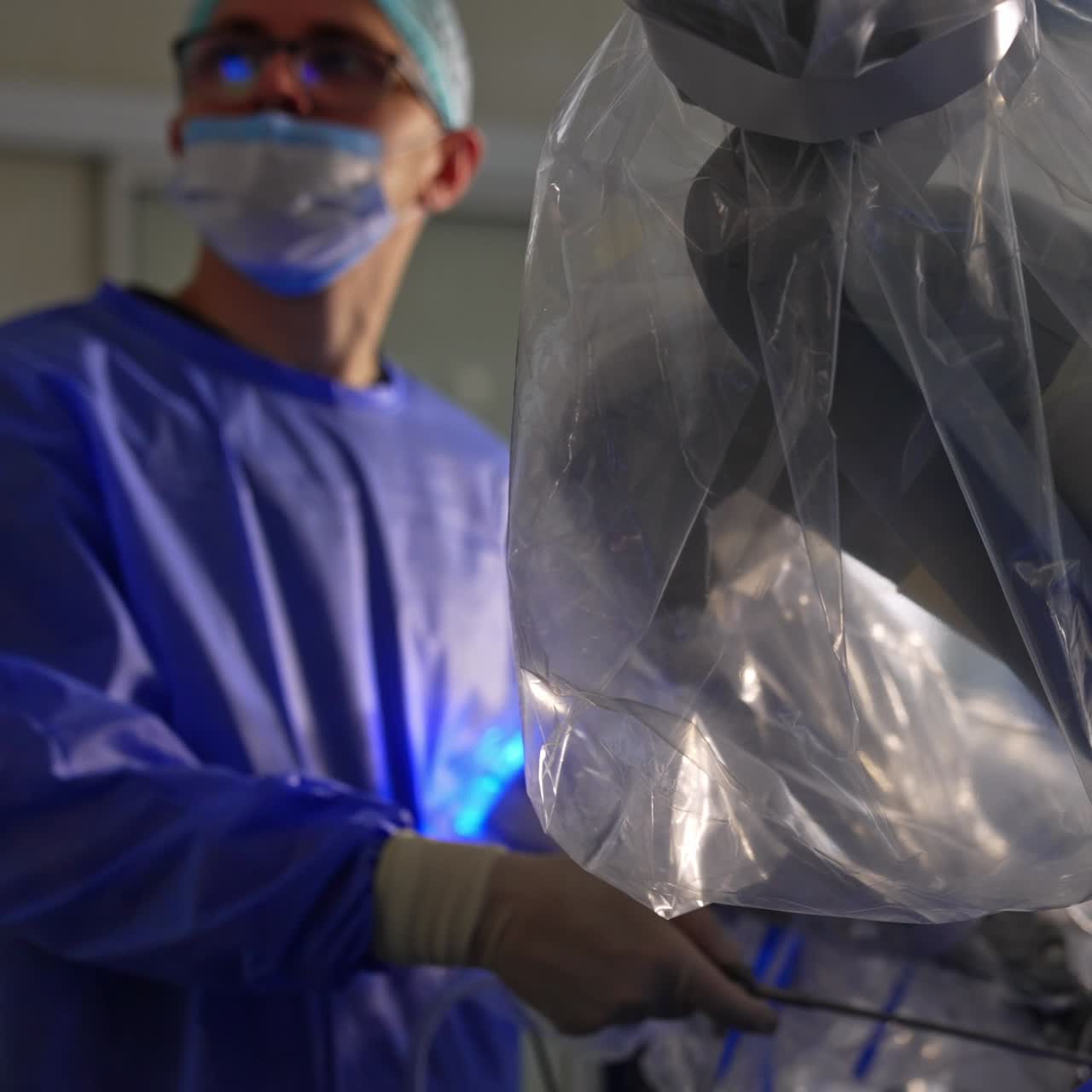 Big robotic arm covered in plastic conducts laparoscopy to the patient. Surgeon standing beside with instrument in his hand looks on the screen