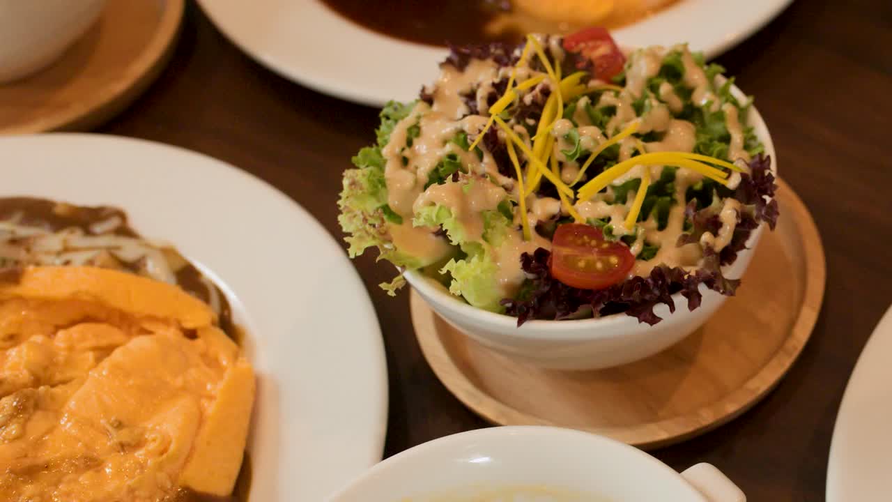 Camera pans over Japanese omelette rice, hamburger steak, salad, and soup in warm restaurant lighting