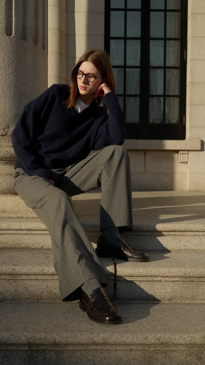 Vertical shot of young female model wearing glasses sweater and trousers sitting on stone palace stairs at sunset