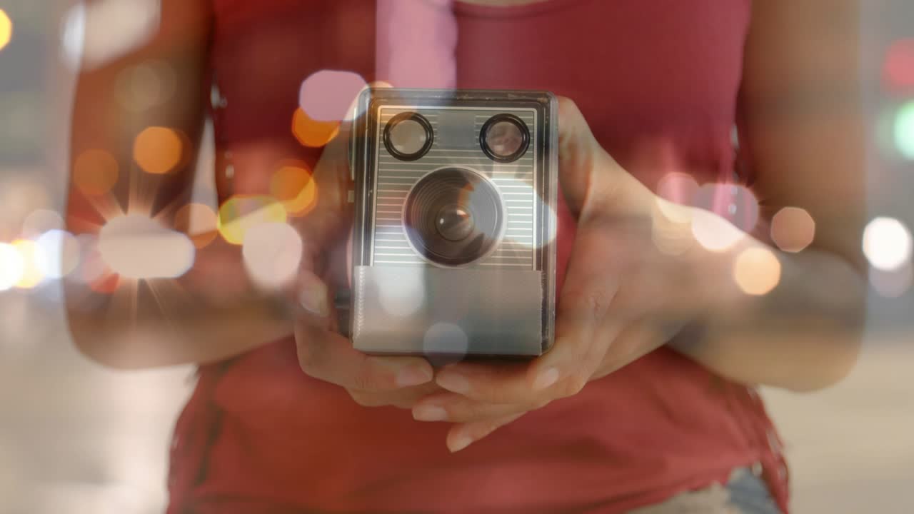 Fotografía que muestra una sonrisa dibujándose, revelando a una mujer con blusa roja sujetando una cámara entre luces bokeh.