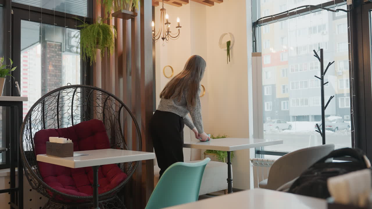 Elegant young woman places lunch on table in stylish indoor cafe surrounded by modern table and chair settings, with large window showing street view of cars passing, flowers and wall mirrors