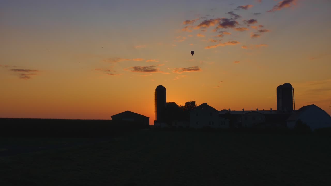 Mongolfiera che si staglia contro un'alba mattutina con fienili e silos in silhouette mentre attraversa il cielo in una mattina d'estate