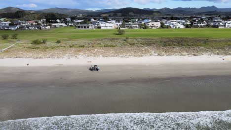 golf cart driving on white sand beach of matarangi in waikato, new zealand with beach houses on the coastline