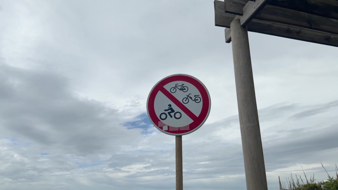 曇り空の下、木造のシェルターの横に、自転車、原動機付自転車、オートバイの乗り入れを禁止する標識が立っている。