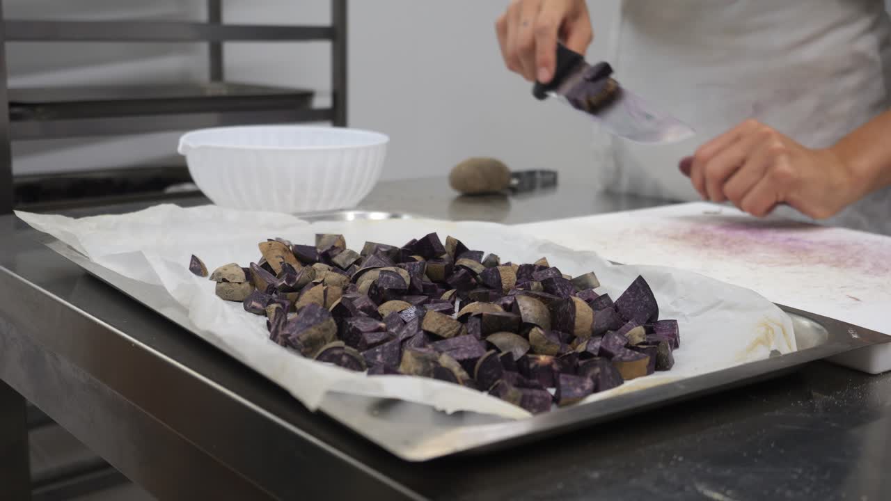 Purple potatoes are chopped on a white cutting board beside a tray lined with parchment paper. A whole potato and bowl sit nearby, with a chef’s hands actively preparing ingredients in pro kitchen set