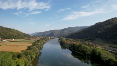 the drina river flows serenely through a lush valley, surrounded by expansive green hills and cultivated farmland, under a clear blue sky with light clouds drifting above