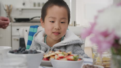 cute-little-asian-boy-eating-fresh-waffles-for-breakfast-enjoying-delicious-homemade-meal-with-family-in-kitchen-at-home-4k