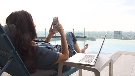 side view of asian woman relaxing peacefully in a chair on skyscraper near swimming pool in bright sunlight using laptop and choosing music favorite on app smartphone and blurred background city.