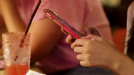 A young woman scrolls on her smartphone with one hand while seated at a table with a pink drink, under warm ambient lighting in a casual Bangkok eatery