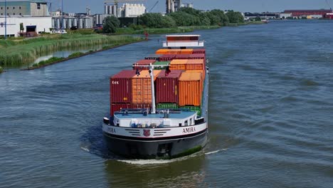 un buque de carga amira que transporta contenedores en un río con edificios de la ciudad en el fondo