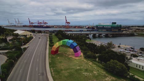 aerial drone backward moving shot over marcus canning’s ‘rainbow’ arch made of containers, container ship docked along port beach, north fremantle, west australia on a cloudy day