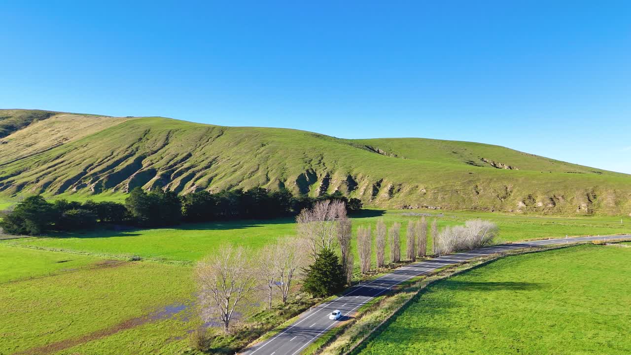 A car travels along a winding road through lush green hills under clear blue skies in Akaroa, New Zealand