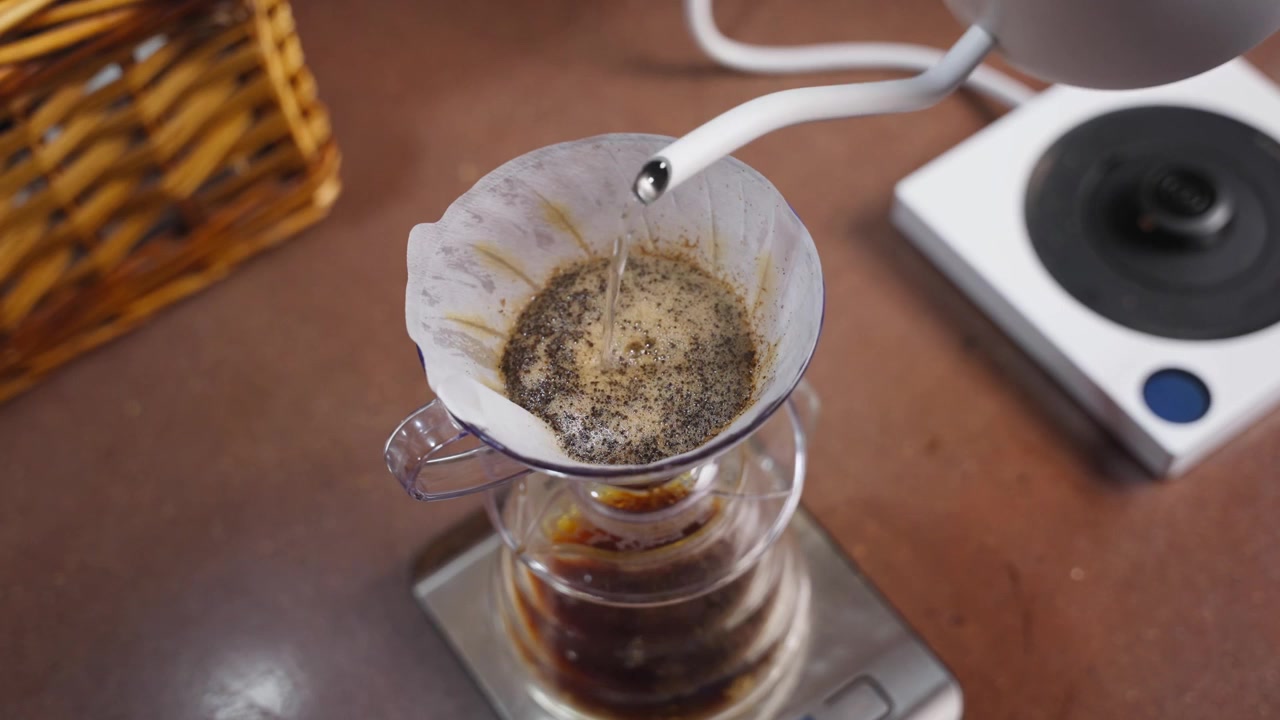 Close-up dari atas penyeduhan kopi pour-over melalui filter kertas, dengan cerat ketel, bubuk kopi yang mengembang, dripper kaca, dan suasana persiapan kafe yang nyaman