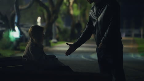 shady handoff in a dark park — a woman on a bench discreetly passes an envelope to a standing man. nighttime secret meeting with suspenseful, criminal or espionage undertones. secret agent word. night