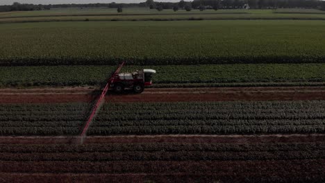 Seguimiento-Aéreo-Lateral-De-Un-Tractor-Que-Arrastra-Amplios-Brazos-Rociadores-Y-Existencias-De-Líquido-En-Los-Alrededores-De-Las-Tierras-De-Cultivo-Con-Varios-Cultivos-En-Segundo-Plano-Y-En-Primer-Plano