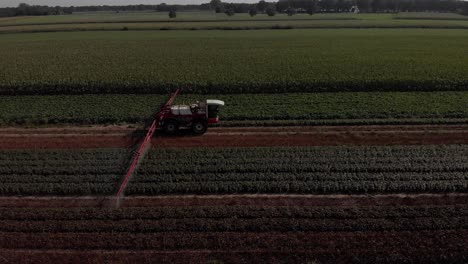 seguimiento aéreo lateral de un tractor que arrastra amplios brazos rociadores y existencias de líquido en los alrededores de las tierras de cultivo con varios cultivos en segundo plano y en primer plano