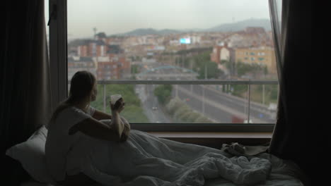 woman in bed drinking tea and looking at city